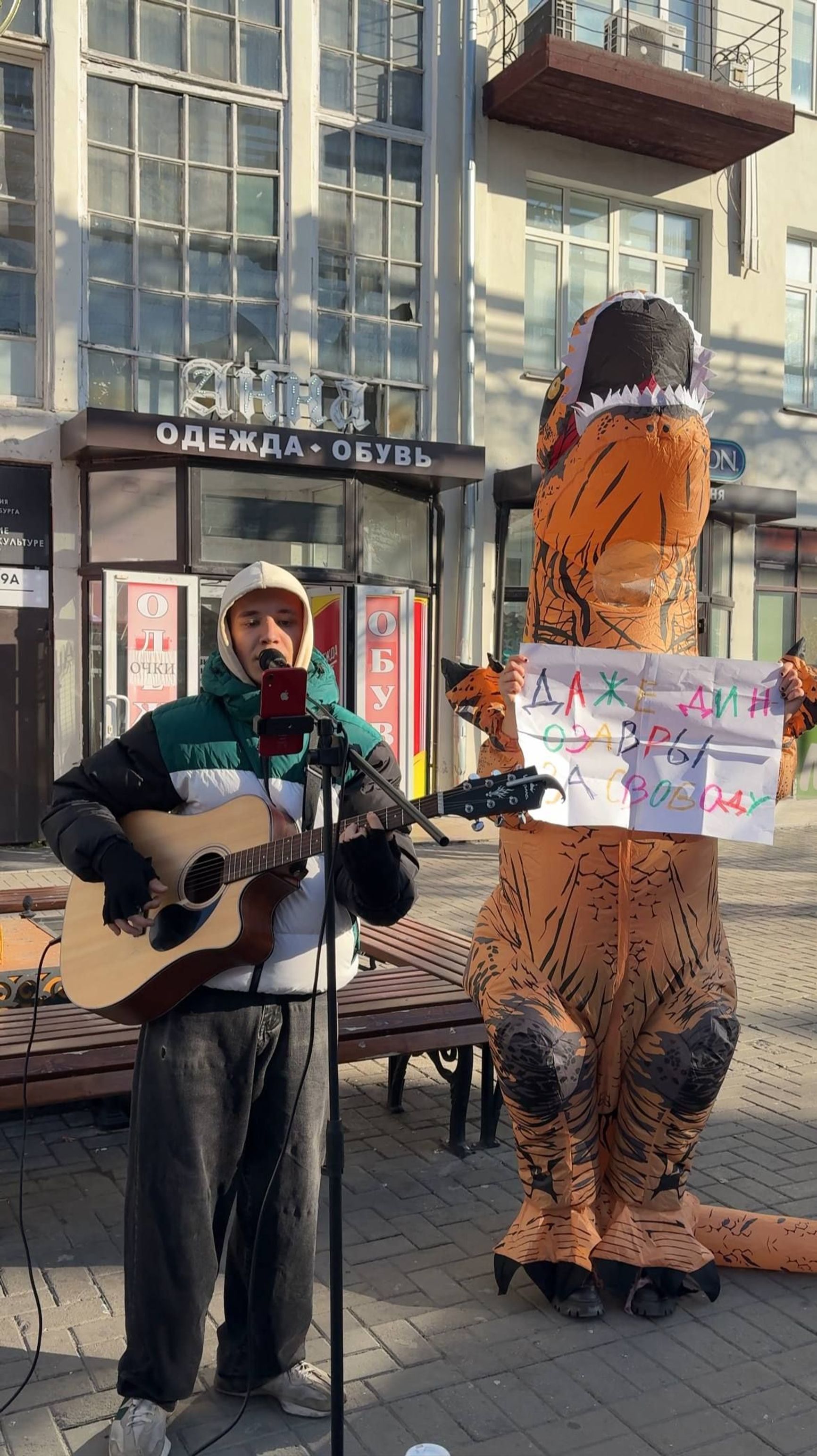Yevgeny Mikhailov (Zhenka Radost) performing next to a person in a dinosaur costume holding a sign that says “Even dinosaurs [support] freedom.”